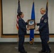 Two female U.S. Air Force personnel are participating in a formal military ceremony, with one officer presenting a wooden plaque to a Senior Master Sergeant. The plaque features the Senior Master Sergeant rank insignia and appears to be a promotion or recognition award. The ceremony takes place in front of U.S. and Air Force flags in what appears to be a conference or briefing room.