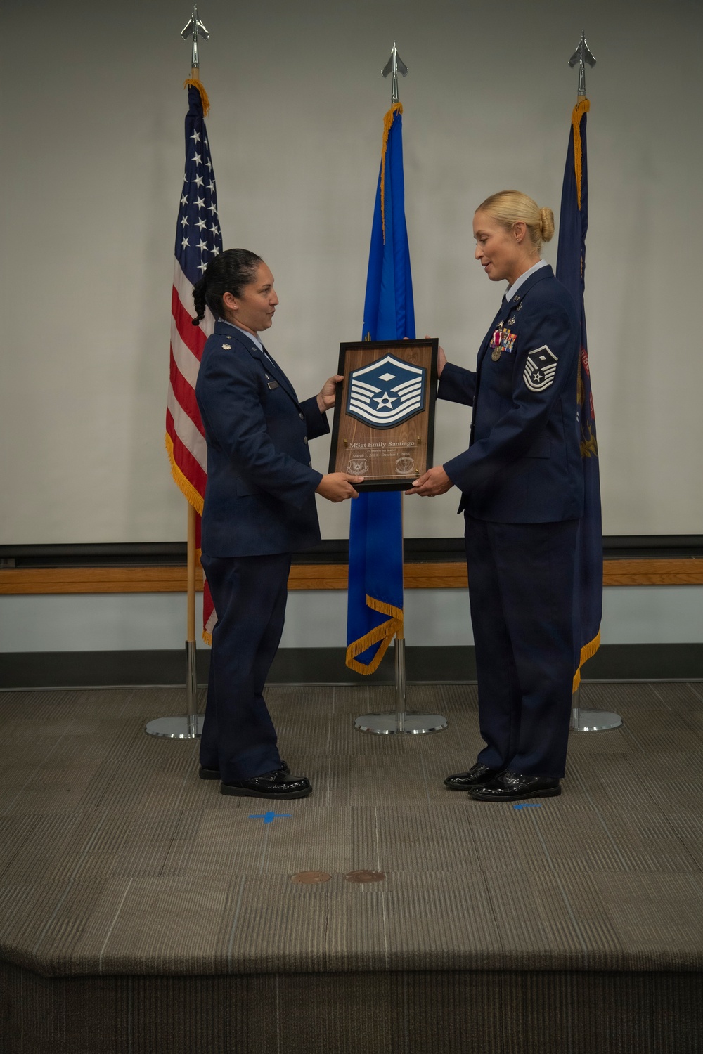 Two U.S. Air Force female personnel in service dress uniforms are on a stage conducting a military promotion or recognition ceremony. The individual on the right is receiving a wooden shadow box plaque bearing a Master Sergeant chevron insignia, with text reading 'MSgt Emily Santiago' visible on the plaque. Three flags — the U.S. flag, Air Force flag, and what appears to be a command or unit flag — are displayed in the background.