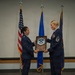 Two U.S. Air Force female personnel in service dress uniforms are on a stage conducting a military promotion or recognition ceremony. The individual on the right is receiving a wooden shadow box plaque bearing a Master Sergeant chevron insignia, with text reading 'MSgt Emily Santiago' visible on the plaque. Three flags — the U.S. flag, Air Force flag, and what appears to be a command or unit flag — are displayed in the background.