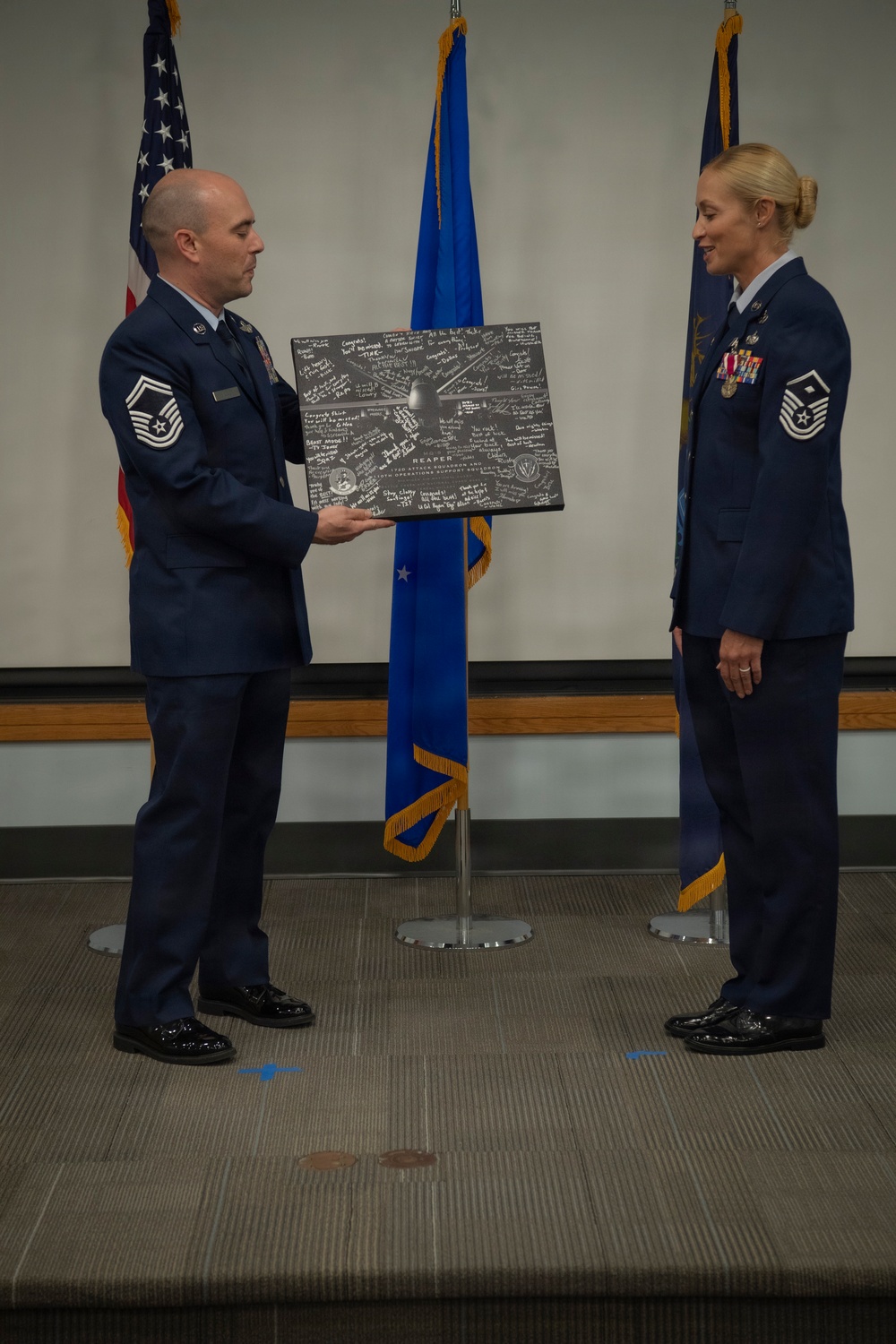 Two U.S. Air Force personnel in dress blue uniforms are participating in a formal military ceremony on a stage. A male Senior Master Sergeant is presenting a signed commemorative plaque to a female Senior Master Sergeant, who is wearing service ribbons and medals. Three flags — the U.S. flag, Air Force flag, and what appears to be a unit flag — are displayed in the background.