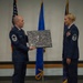 Two U.S. Air Force personnel in dress blue uniforms are participating in a formal military ceremony on a stage. A male Senior Master Sergeant is presenting a signed commemorative plaque to a female Senior Master Sergeant, who is wearing service ribbons and medals. Three flags — the U.S. flag, Air Force flag, and what appears to be a unit flag — are displayed in the background.