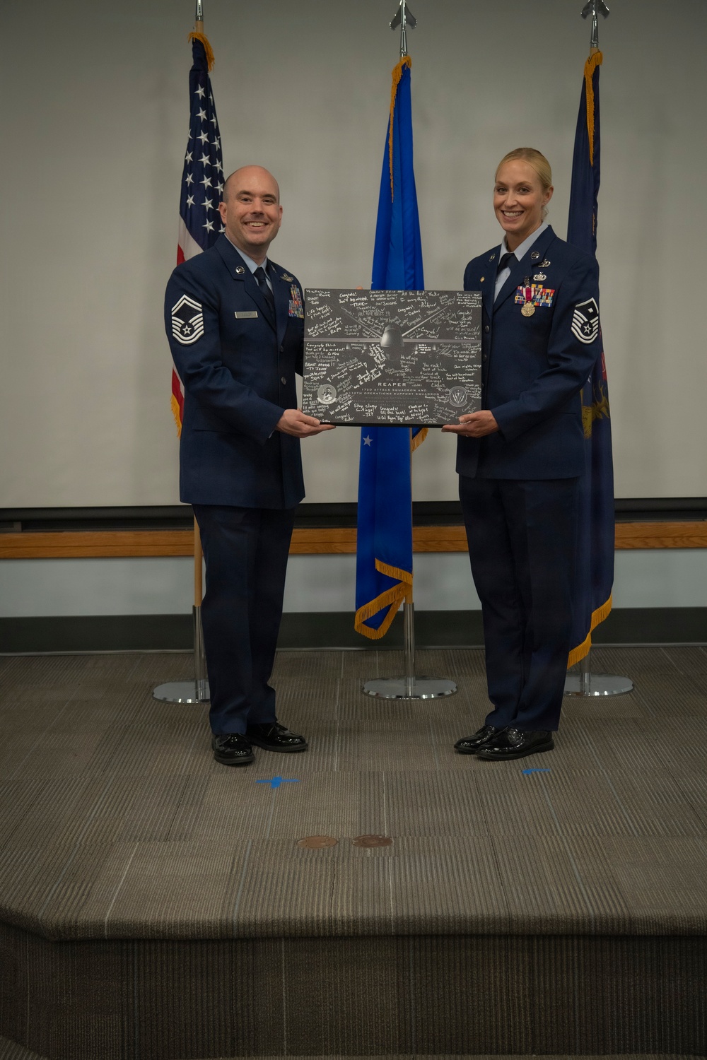 Two U.S. Air Force personnel in dress blue uniforms are posing together on a stage, jointly holding a commemorative plaque or shadow box covered in signatures. The item appears to reference a 'REAPER' aircraft and includes unit insignia, suggesting a retirement, promotion, or farewell ceremony. Three flags — the U.S. flag and two Air Force flags — are displayed in the background.