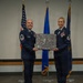 Two U.S. Air Force personnel in dress blue uniforms are posing together on a stage, jointly holding a commemorative plaque or shadow box covered in signatures. The item appears to reference a 'REAPER' aircraft and includes unit insignia, suggesting a retirement, promotion, or farewell ceremony. Three flags — the U.S. flag and two Air Force flags — are displayed in the background.