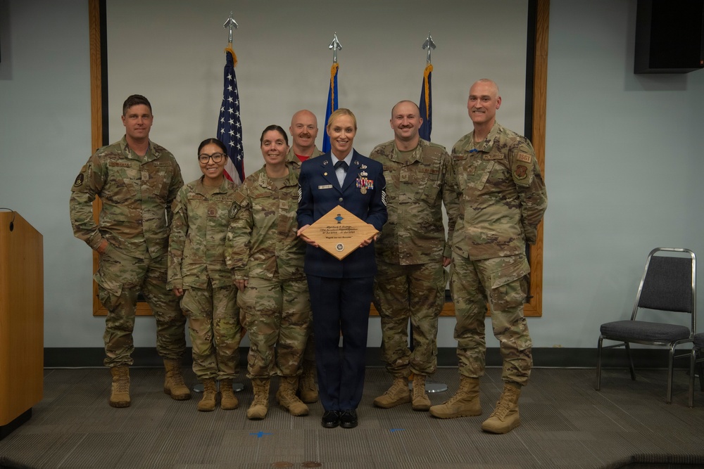 A group of seven military personnel pose for a formal photo in what appears to be a ceremony room. The central figure, wearing a dress blue Air Force uniform, holds a wooden plaque award while the others wear Army Combat Uniform (ACU/OCP camouflage pattern). This appears to be a military recognition or retirement ceremony.