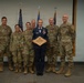 A group of seven military personnel pose for a formal photo in what appears to be a ceremony room. The central figure, wearing a dress blue Air Force uniform, holds a wooden plaque award while the others wear Army Combat Uniform (ACU/OCP camouflage pattern). This appears to be a military recognition or retirement ceremony.