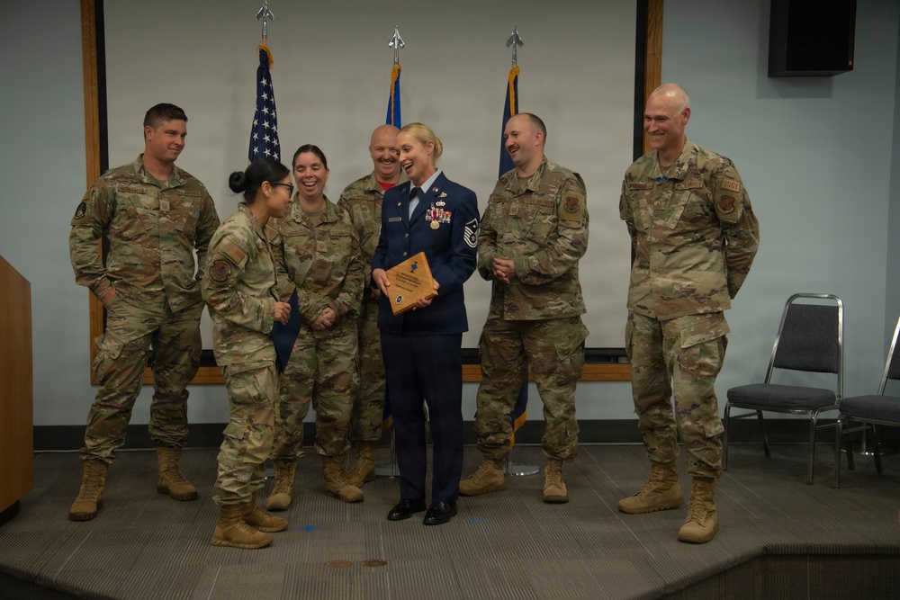 A military award or recognition ceremony is taking place in what appears to be a conference or briefing room. A female service member in dress blues uniform is holding a wooden plaque/award while surrounded by six colleagues in OCP (Operational Camouflage Pattern) uniforms. The group is smiling and laughing, suggesting a celebratory atmosphere.