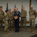 A military award or recognition ceremony is taking place in what appears to be a conference or briefing room. A female service member in dress blues uniform is holding a wooden plaque/award while surrounded by six colleagues in OCP (Operational Camouflage Pattern) uniforms. The group is smiling and laughing, suggesting a celebratory atmosphere.