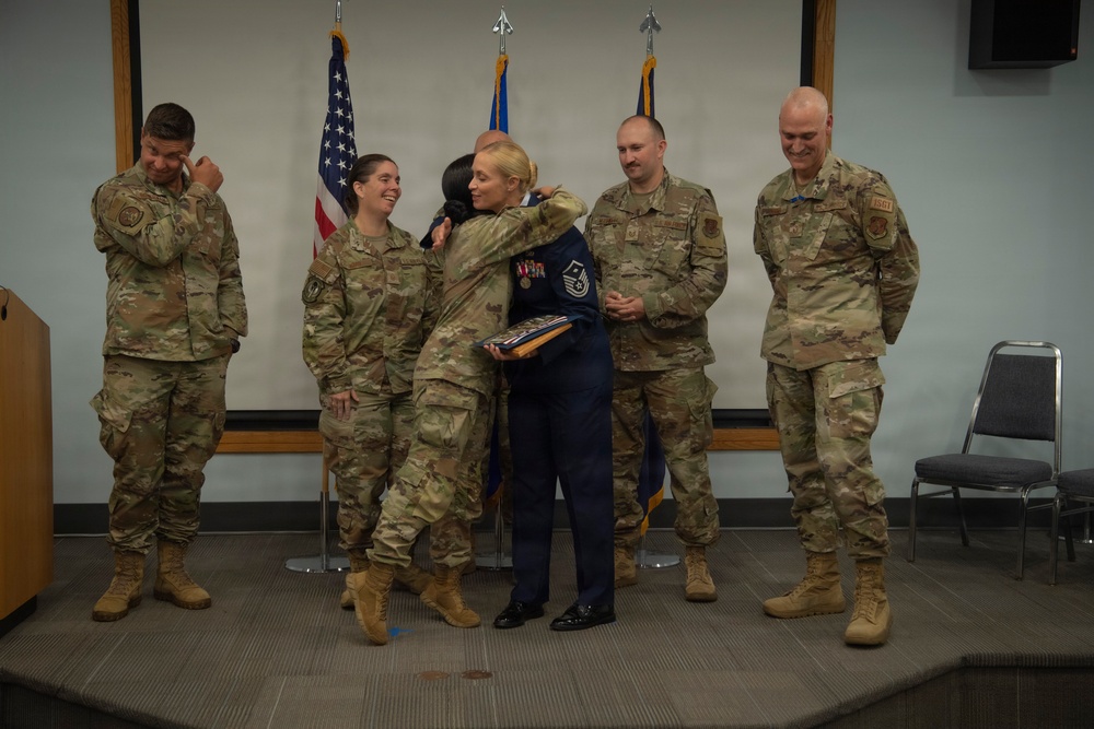 A military ceremony is taking place on a small stage, likely a retirement or award ceremony. A service member in dress blues is embracing another person while holding a folded flag or certificate, surrounded by colleagues in OCP (Operational Camouflage Pattern) uniforms. The atmosphere appears celebratory and emotional, with onlookers smiling and one person appearing to wipe away tears.