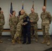 A military ceremony is taking place on a small stage, likely a retirement or award ceremony. A service member in dress blues is embracing another person while holding a folded flag or certificate, surrounded by colleagues in OCP (Operational Camouflage Pattern) uniforms. The atmosphere appears celebratory and emotional, with onlookers smiling and one person appearing to wipe away tears.