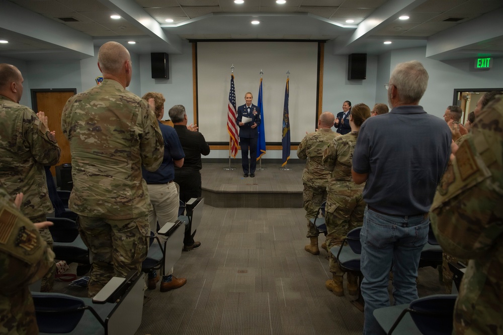 A military ceremony is taking place in an indoor briefing or conference room, where a female officer in dress blue uniform stands at a podium on a small stage flanked by the American flag and what appears to be a U.S. Air Force flag. The audience, consisting of military personnel in OCP (Operational Camouflage Pattern) uniforms and some civilians, are standing and applauding, suggesting a promotion, retirement, or award ceremony. A second uniformed individual stands to the right of the central figure on the stage.