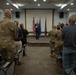 A military ceremony is taking place in an indoor briefing or conference room, where a female officer in dress blue uniform stands at a podium on a small stage flanked by the American flag and what appears to be a U.S. Air Force flag. The audience, consisting of military personnel in OCP (Operational Camouflage Pattern) uniforms and some civilians, are standing and applauding, suggesting a promotion, retirement, or award ceremony. A second uniformed individual stands to the right of the central figure on the stage.