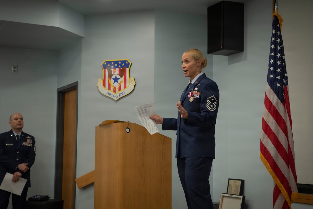 A female U.S. Air Force senior non-commissioned officer is speaking at a podium during an official military ceremony at the 110th Wing. She is in dress blues and holds papers while addressing an audience. A male officer in dress blues stands in the background to the left.
