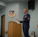 A female U.S. Air Force senior non-commissioned officer is speaking at a podium during an official military ceremony at the 110th Wing. She is in dress blues and holds papers while addressing an audience. A male officer in dress blues stands in the background to the left.