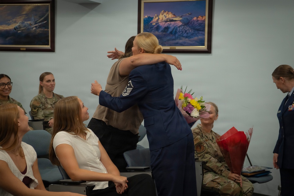 A military ceremony appears to be taking place in a conference or briefing room, where a female Air Force Master Sergeant in dress blues is embracing a civilian woman. Several attendees are seated, including personnel in military camouflage uniforms, and flowers are visible being held by a seated service member, suggesting a promotion, retirement, or recognition ceremony.