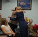 A military ceremony appears to be taking place in a conference or briefing room, where a female Air Force Master Sergeant in dress blues is embracing a civilian woman. Several attendees are seated, including personnel in military camouflage uniforms, and flowers are visible being held by a seated service member, suggesting a promotion, retirement, or recognition ceremony.