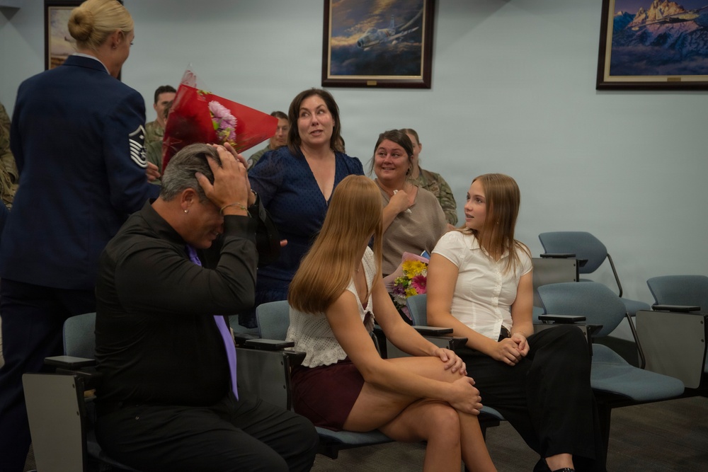 A military ceremony appears to be taking place in what looks like a briefing or meeting room, with a uniformed service member presenting flowers to a group of civilians. Several people display strong emotional reactions including surprise and tears, suggesting this may be a recognition, retirement, or homecoming event. Two young women are seated in the foreground while adults behind them react with visible emotion.