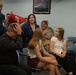 A military ceremony appears to be taking place in what looks like a briefing or meeting room, with a uniformed service member presenting flowers to a group of civilians. Several people display strong emotional reactions including surprise and tears, suggesting this may be a recognition, retirement, or homecoming event. Two young women are seated in the foreground while adults behind them react with visible emotion.