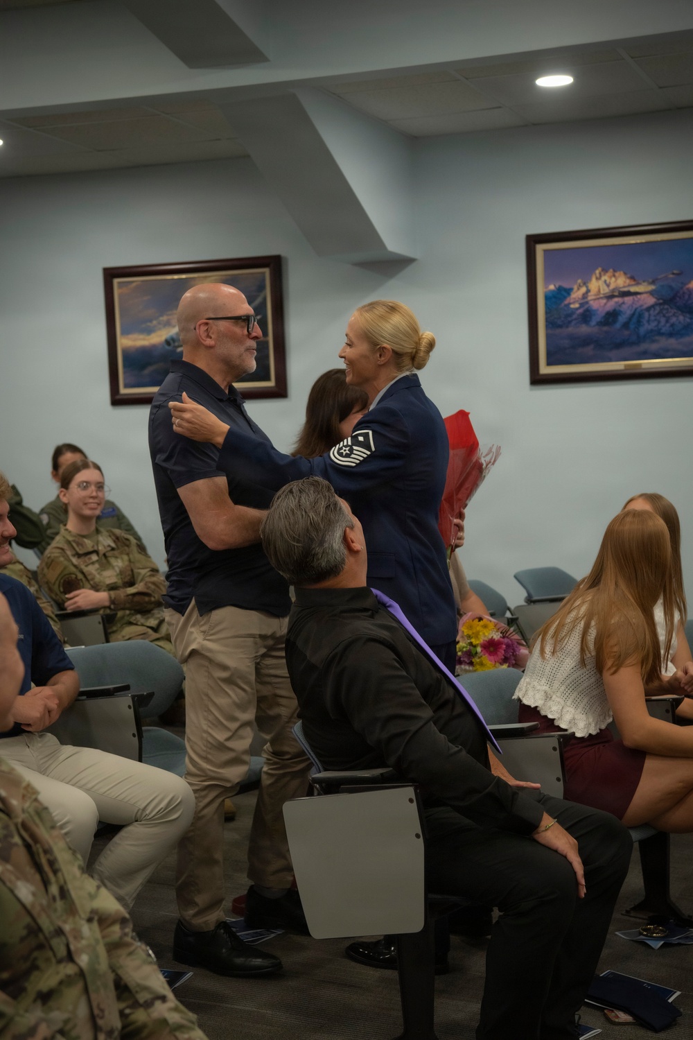 A military ceremony appears to be taking place in a conference or briefing room, likely a promotion or retirement event. A female U.S. Air Force senior non-commissioned officer, wearing a service dress uniform with Senior Master Sergeant chevrons, is embracing a civilian man while holding a bouquet of red flowers. Attendees including uniformed military personnel and civilians are seated and observing the moment.