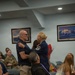 A military ceremony appears to be taking place in a conference or briefing room, likely a promotion or retirement event. A female U.S. Air Force senior non-commissioned officer, wearing a service dress uniform with Senior Master Sergeant chevrons, is embracing a civilian man while holding a bouquet of red flowers. Attendees including uniformed military personnel and civilians are seated and observing the moment.