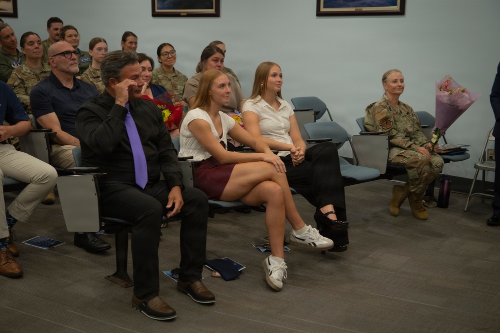 A formal military ceremony is taking place in an indoor venue, with a mixed audience of uniformed military personnel and civilian attendees seated in rows. Several people in the front rows appear emotionally moved, with one man wiping his eye, suggesting a significant milestone event such as a promotion, retirement, or graduation ceremony. Flowers are visible being held by attendees, reinforcing the celebratory nature of the occasion.