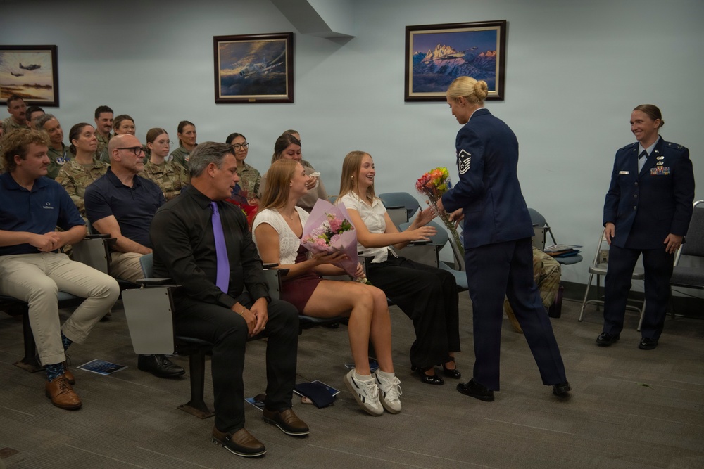 A military ceremony is taking place in a formal meeting room, where a female Air Force Master Sergeant in dress blues is presenting flowers to a young woman seated in the audience. The room is filled with a mix of uniformed military personnel and civilian attendees, suggesting a promotion or retirement ceremony. A second female officer in dress blues stands nearby smiling, observing the presentation.