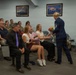 A military ceremony is taking place in a formal meeting room, where a female Air Force Master Sergeant in dress blues is presenting flowers to a young woman seated in the audience. The room is filled with a mix of uniformed military personnel and civilian attendees, suggesting a promotion or retirement ceremony. A second female officer in dress blues stands nearby smiling, observing the presentation.