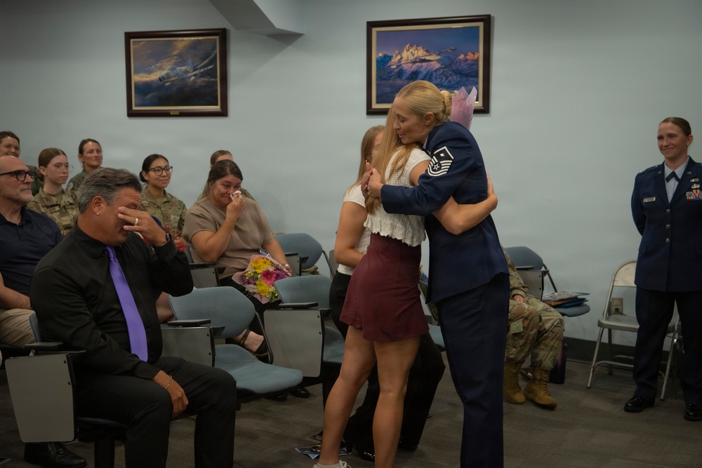 A military promotion or retirement ceremony is taking place in what appears to be a U.S. Air Force facility. A female Airman in dress blues, bearing Master Sergeant chevrons, is embracing a young woman while emotional family members and colleagues look on. The moment is clearly significant, with a man covering his face in tears and a woman wiping her eyes in the background.