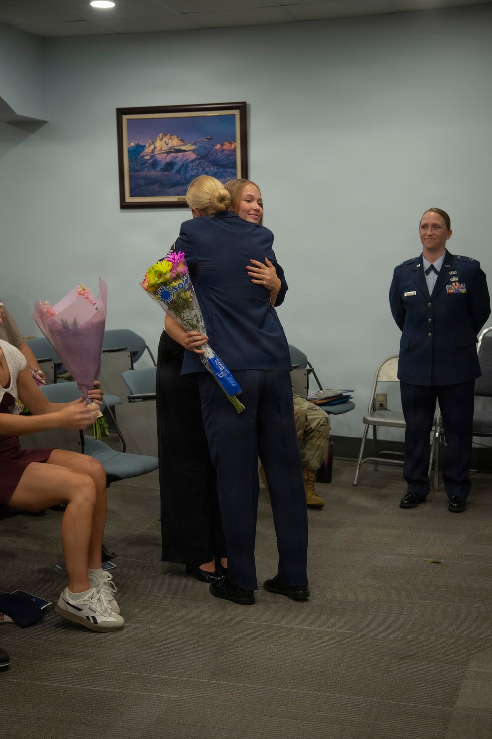 A military ceremony appears to be taking place in a small indoor room, likely a US Air Force facility. Two individuals in dark blue uniforms are embracing while one holds a bouquet of flowers, suggesting a recognition or promotion event. A uniformed US Air Force officer with rank insignia and ribbons stands observing in the background.