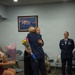 A military ceremony appears to be taking place in a small indoor room, likely a US Air Force facility. Two individuals in dark blue uniforms are embracing while one holds a bouquet of flowers, suggesting a recognition or promotion event. A uniformed US Air Force officer with rank insignia and ribbons stands observing in the background.
