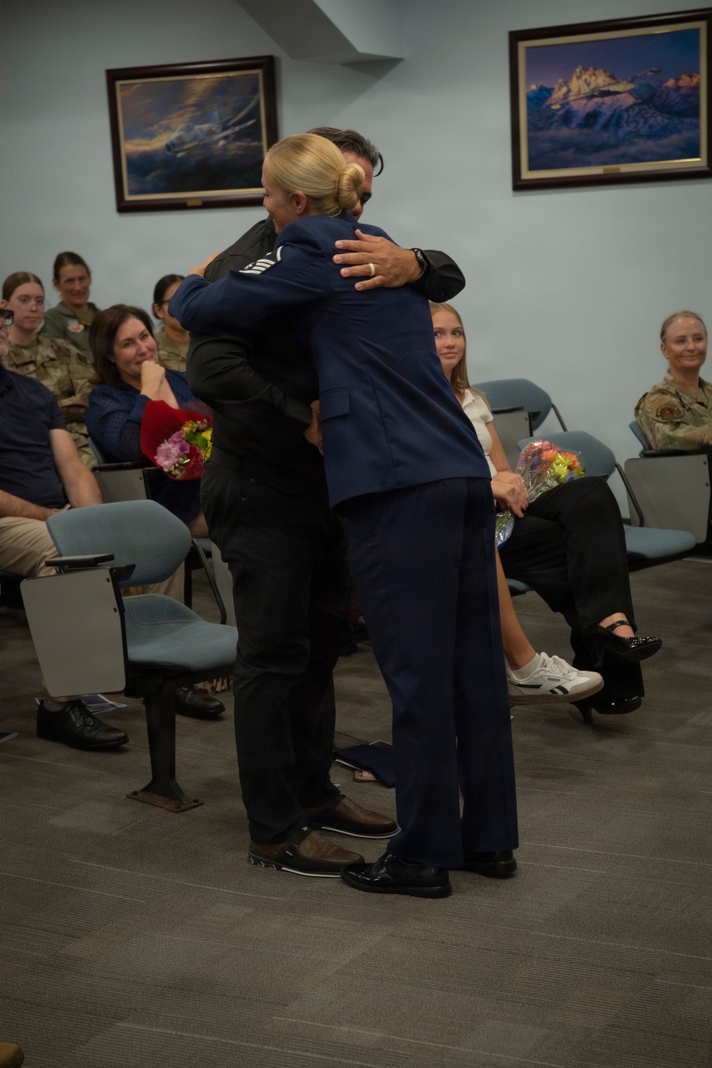 A military ceremony taking place in what appears to be a briefing or conference room on a military installation. A person in a U.S. Air Force dress blue uniform is embracing a civilian in the foreground, while seated audience members — including personnel in military camouflage uniforms and civilians holding flowers — look on with smiles. The setting and flowers suggest a promotion, retirement, or similar formal recognition ceremony.