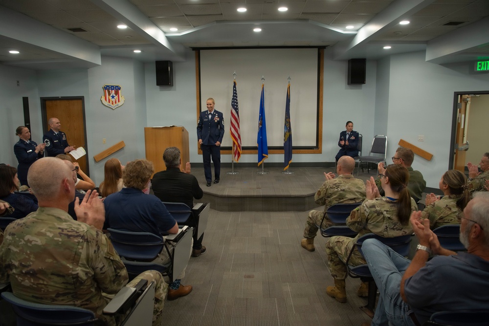 A military ceremony is taking place in a formal briefing room, with a uniformed officer standing at the front of a small stage receiving applause from an audience of military personnel and civilians. The setting includes an American flag and two blue flags flanking a wooden podium, along with a military unit insignia mounted on the wall. The atmosphere suggests a promotion, award, or change-of-command ceremony.