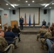 A military ceremony is taking place in a formal briefing room, with a uniformed officer standing at the front of a small stage receiving applause from an audience of military personnel and civilians. The setting includes an American flag and two blue flags flanking a wooden podium, along with a military unit insignia mounted on the wall. The atmosphere suggests a promotion, award, or change-of-command ceremony.