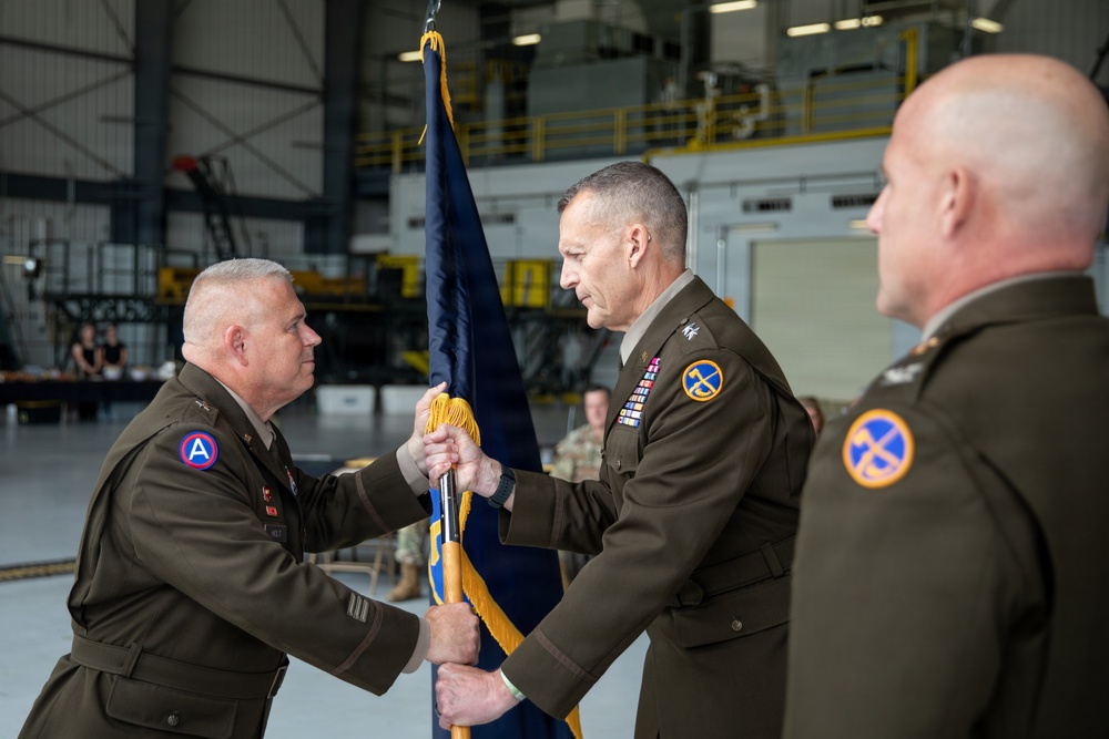 A U.S. Army change of command ceremony is taking place inside a military hangar. Two senior officers are exchanging a unit guidon (flag), which is the traditional symbolic act of transferring command authority. A third officer stands to the right observing the ceremony.