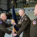 A U.S. Army change of command ceremony is taking place inside a military hangar. Two senior officers are exchanging a unit guidon (flag), which is the traditional symbolic act of transferring command authority. A third officer stands to the right observing the ceremony.