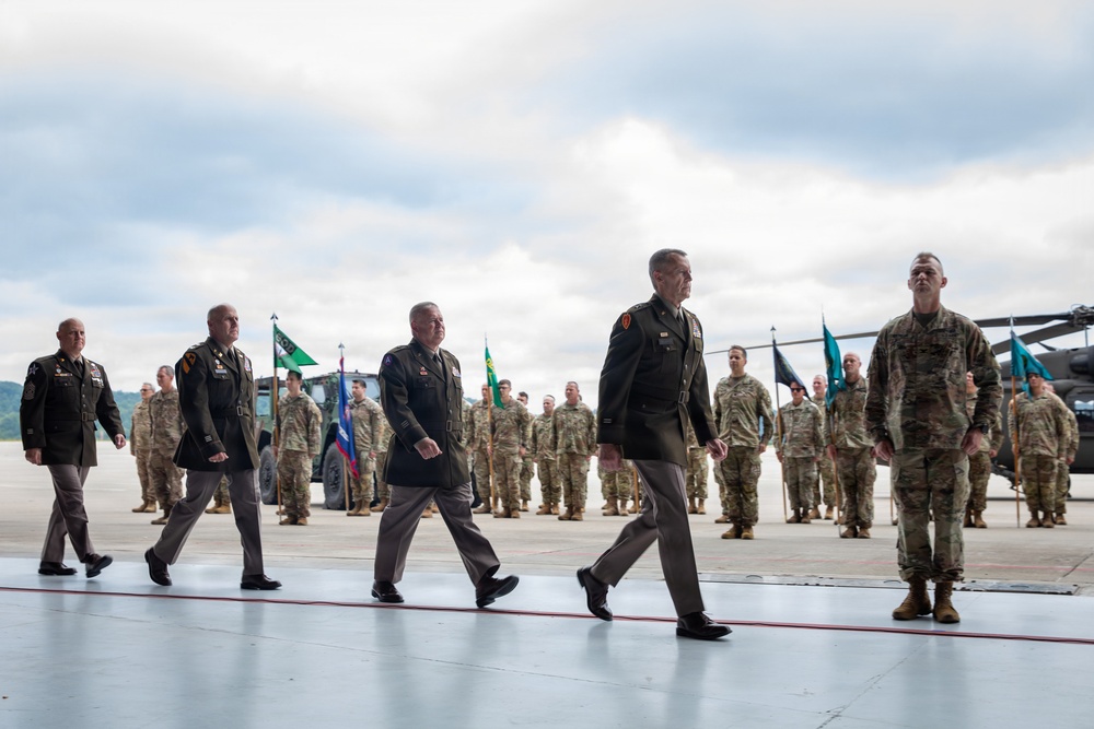 A formal military ceremony is taking place on what appears to be an airfield or tarmac. Senior officers in dress uniforms are walking in procession while troops in combat uniforms stand in formation behind them. Military flags and guidons are visible in the background alongside what appears to be a military helicopter.