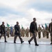 A formal military ceremony is taking place on what appears to be an airfield or tarmac. Senior officers in dress uniforms are walking in procession while troops in combat uniforms stand in formation behind them. Military flags and guidons are visible in the background alongside what appears to be a military helicopter.