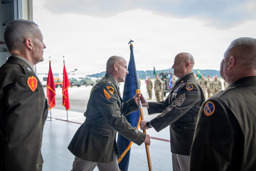 A formal U.S. Army military ceremony, likely a change of command or activation ceremony, taking place on what appears to be an airfield or hangar apron. Two central figures in Army Service Uniforms are exchanging a unit guidon (flag on a staff), which is the traditional symbolic act of transferring command authority. Additional uniformed personnel are present as witnesses, with formation troops visible in the background.