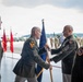 A formal U.S. Army military ceremony, likely a change of command or activation ceremony, taking place on what appears to be an airfield or hangar apron. Two central figures in Army Service Uniforms are exchanging a unit guidon (flag on a staff), which is the traditional symbolic act of transferring command authority. Additional uniformed personnel are present as witnesses, with formation troops visible in the background.