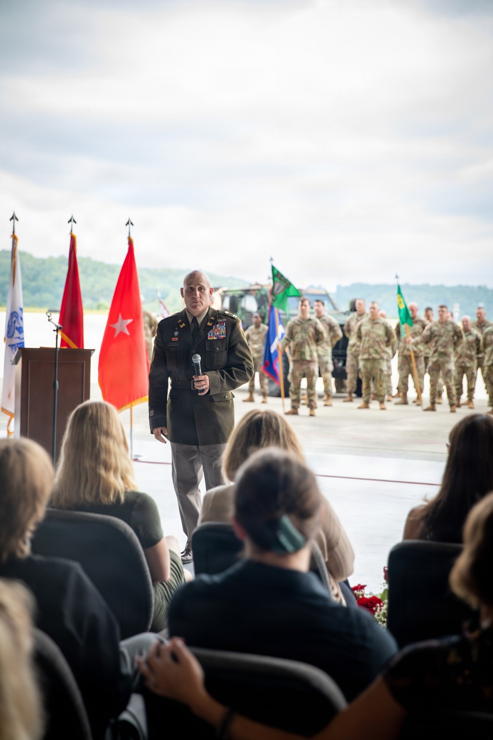 A U.S. Army officer in dress green uniform is addressing an audience at what appears to be a military ceremony, likely a change of command or unit activation event. He is holding a microphone and standing before a formation of soldiers in Army Combat Uniforms, with multiple military flags and guidons visible in the background. Civilian guests are seated in the foreground facing the speaker.