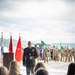 A U.S. Army officer in dress green uniform is addressing an audience at what appears to be a military ceremony, likely a change of command or unit activation event. He is holding a microphone and standing before a formation of soldiers in Army Combat Uniforms, with multiple military flags and guidons visible in the background. Civilian guests are seated in the foreground facing the speaker.