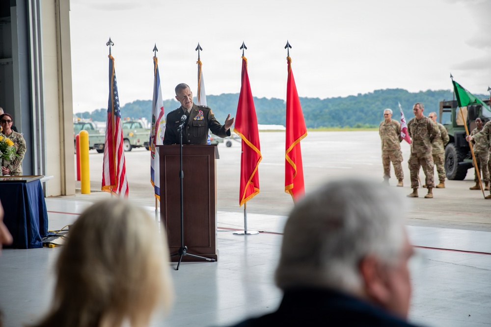A U.S. Army officer in dress uniform delivers a speech at a podium during a formal military ceremony held inside or at the entrance of an aircraft hangar. Multiple flags, including the U.S. flag and red Army unit colors, are displayed behind the speaker. Uniformed soldiers and civilians are present as an audience.