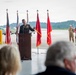 A U.S. Army officer in dress uniform delivers a speech at a podium during a formal military ceremony held inside or at the entrance of an aircraft hangar. Multiple flags, including the U.S. flag and red Army unit colors, are displayed behind the speaker. Uniformed soldiers and civilians are present as an audience.