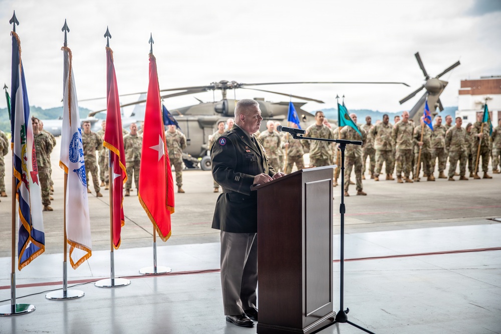 A senior U.S. Army officer is delivering a speech at a podium during a formal military ceremony on what appears to be a flight line or tarmac. Multiple ceremonial flags are displayed to his left, and a large formation of soldiers in Army Combat Uniforms stands at attention in the background. A military helicopter, likely a UH-60 Black Hawk, is visible behind the formation.