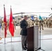A senior U.S. Army officer is delivering a speech at a podium during a formal military ceremony on what appears to be a flight line or tarmac. Multiple ceremonial flags are displayed to his left, and a large formation of soldiers in Army Combat Uniforms stands at attention in the background. A military helicopter, likely a UH-60 Black Hawk, is visible behind the formation.