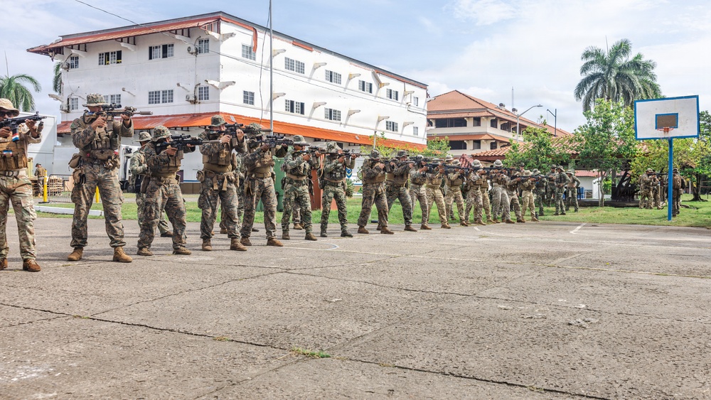 A line of approximately 20 military personnel in camouflage uniforms are conducting a weapons training drill on an outdoor concrete courtyard. They are all in a firing stance with rifles raised and aimed in the same direction. The setting appears to be a military installation in a tropical region, evidenced by palm trees and colonial-style white buildings in the background.