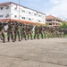 A line of approximately 20 military personnel in camouflage uniforms are conducting a weapons training drill on an outdoor concrete courtyard. They are all in a firing stance with rifles raised and aimed in the same direction. The setting appears to be a military installation in a tropical region, evidenced by palm trees and colonial-style white buildings in the background.