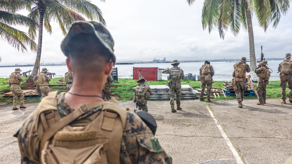 The image shows a group of military personnel conducting what appears to be a tactical training exercise or operation near a waterfront area with palm trees. Multiple soldiers in camouflage uniforms are visible, with some wearing patches labeled 'MARINA' (indicating Mexican Navy personnel), suggesting a joint multinational military exercise. The setting appears to be a tropical coastal location with a harbor or port visible in the background.
