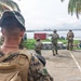 The image shows a group of military personnel conducting what appears to be a tactical training exercise or operation near a waterfront area with palm trees. Multiple soldiers in camouflage uniforms are visible, with some wearing patches labeled 'MARINA' (indicating Mexican Navy personnel), suggesting a joint multinational military exercise. The setting appears to be a tropical coastal location with a harbor or port visible in the background.