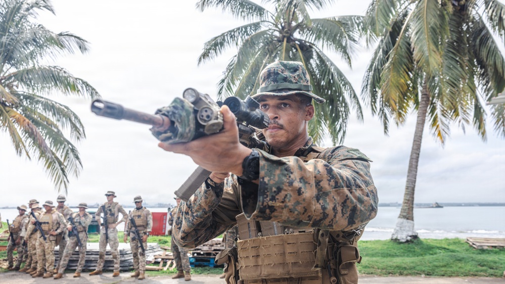 A soldier in MARPAT digital camouflage uniform is aiming a rifle equipped with a scope and tactical light toward the camera in a tropical coastal setting. A group of similarly uniformed personnel stand in the background near palm trees and what appears to be a waterfront area. The scene appears to be a military training exercise or demonstration.