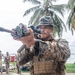 A soldier in MARPAT digital camouflage uniform is aiming a rifle equipped with a scope and tactical light toward the camera in a tropical coastal setting. A group of similarly uniformed personnel stand in the background near palm trees and what appears to be a waterfront area. The scene appears to be a military training exercise or demonstration.
